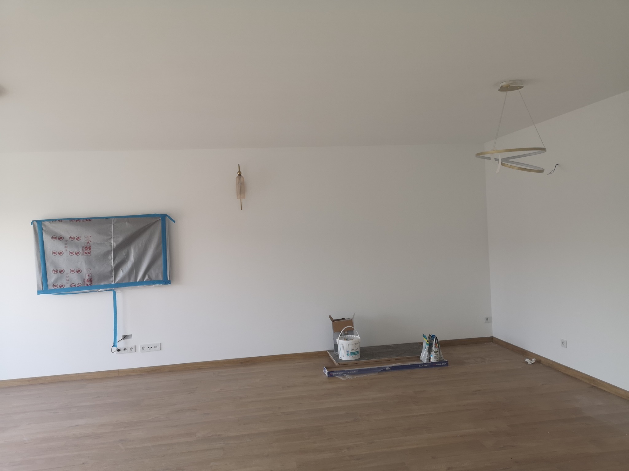 Pokój przed sprzątaniem po remoncie: zabezpieczona folią ściana, puszka farby, listwy przypodłogowe i elementy oświetlenia.
