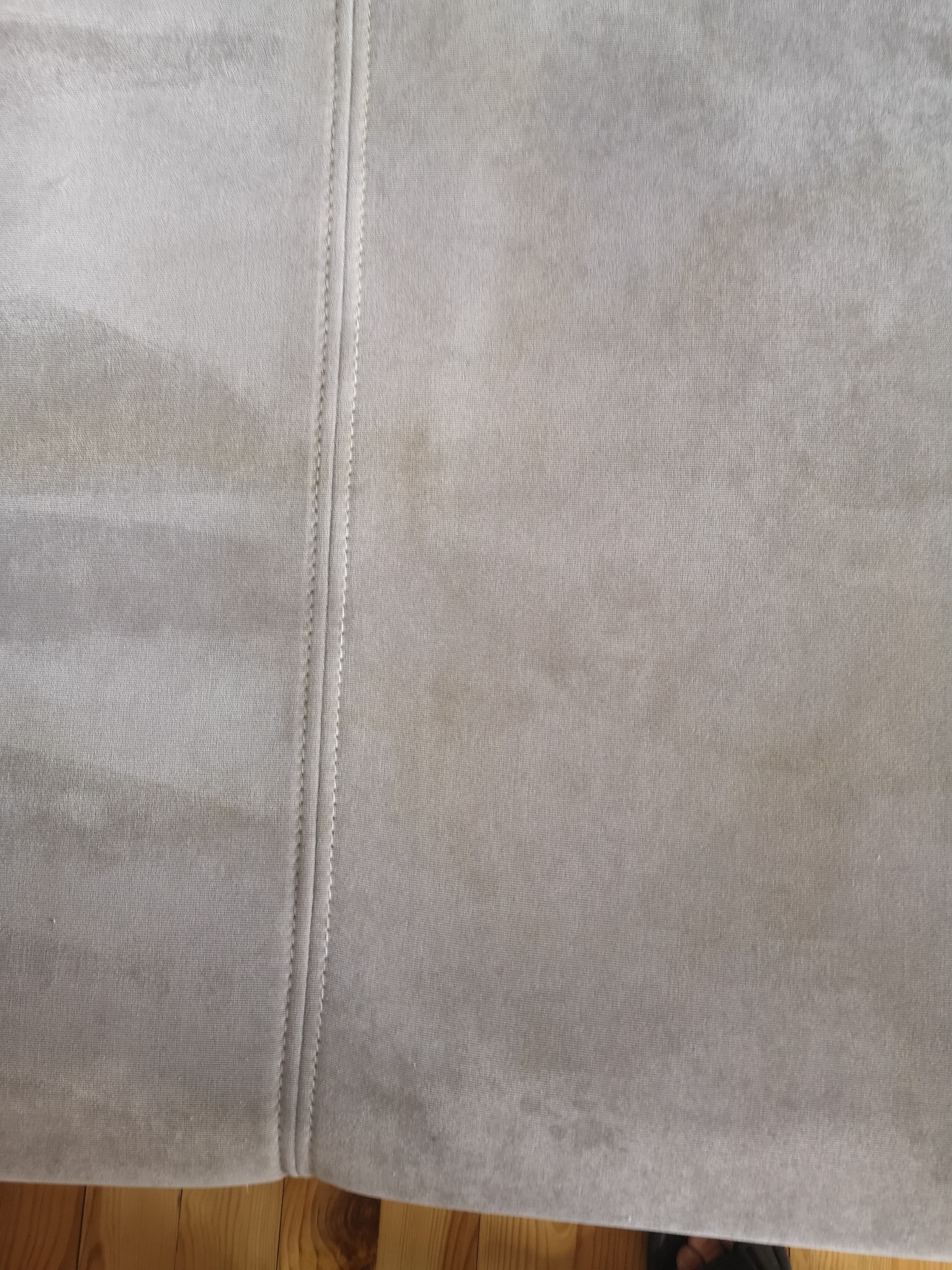 Fragment szarej, welurowej kanapy z widocznym szwem i delikatnymi plamami, częściowo widoczna drewniana podłoga.