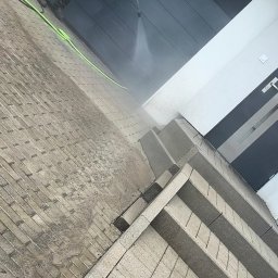 OLEKSANDR MEKHEDA MEKA FASAD - Mężczyzna w kombinezonie myje kostkę brukową przed garażem za pomocą myjki ciśnieniowej. Widoczny efekt czyszczenia, zielony wąż i fragment schodów.