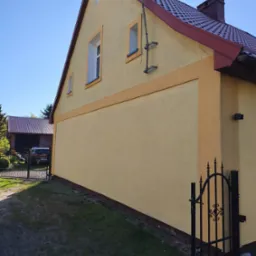 Żółty dom z czerwoną dachówką po malowaniu, widoczna boczna elewacja z oknami i dekoracyjnym paskiem. W tle brama i budynek gospodarczy.