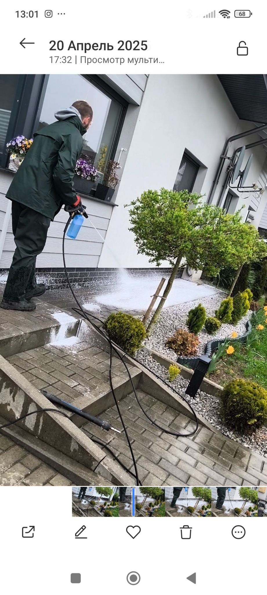 Mężczyzna w zielonym kombinezonie czyści kostkę brukową myjką ciśnieniową. Widoczne rośliny, kwiaty w donicach i elementy elewacji domu. Woda pod ciśnieniem.