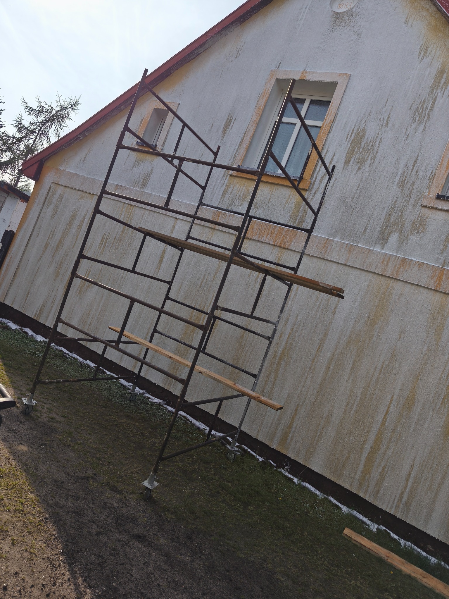 Elewacja budynku przed myciem, widoczne zacieki i rusztowanie. Budynek z oknami w ozdobnych ramach, trawa z resztkami śniegu. Ujęcie z dołu.