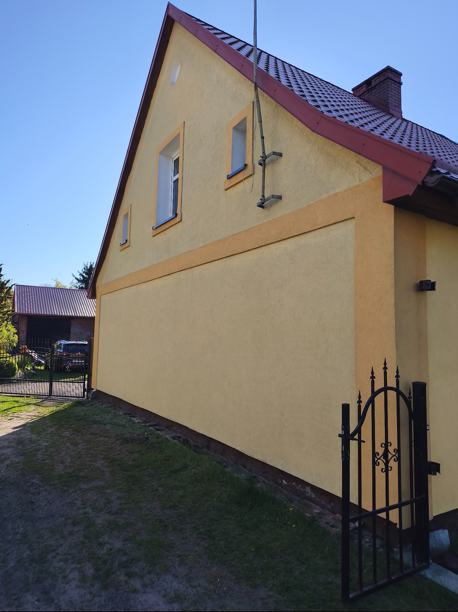 Żółty dom z czerwoną dachówką po malowaniu, widoczna boczna elewacja z oknami i dekoracyjnym paskiem. W tle brama i budynek gospodarczy.