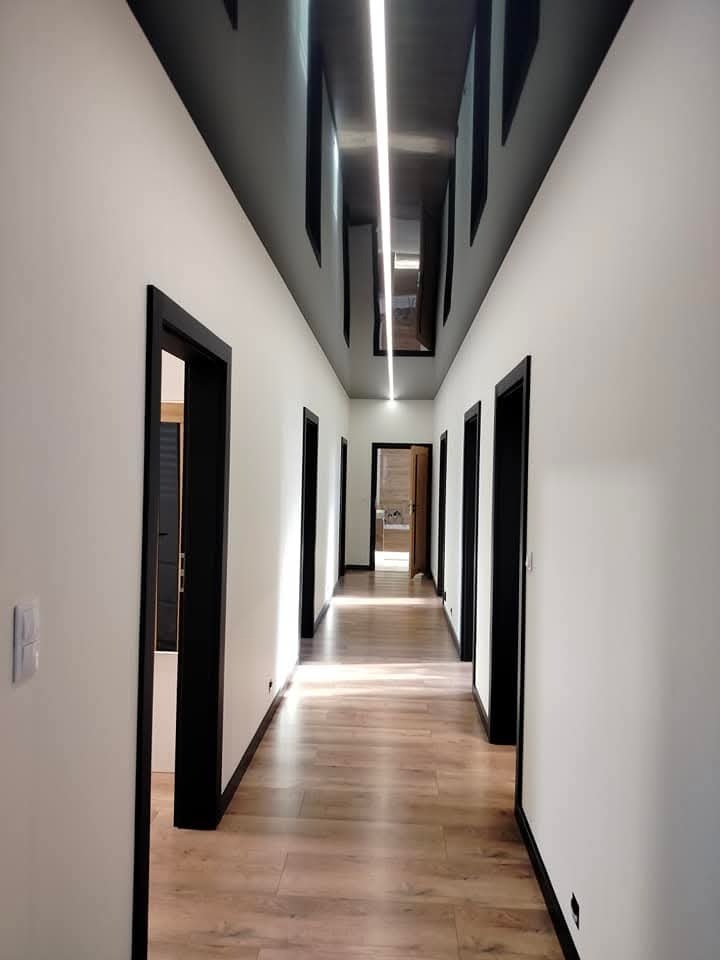 Długi korytarz z panelami podłogowymi, białymi ścianami, czarnymi futrynami drzwi i czarnym sufitem podwieszanym z oświetleniem LED.