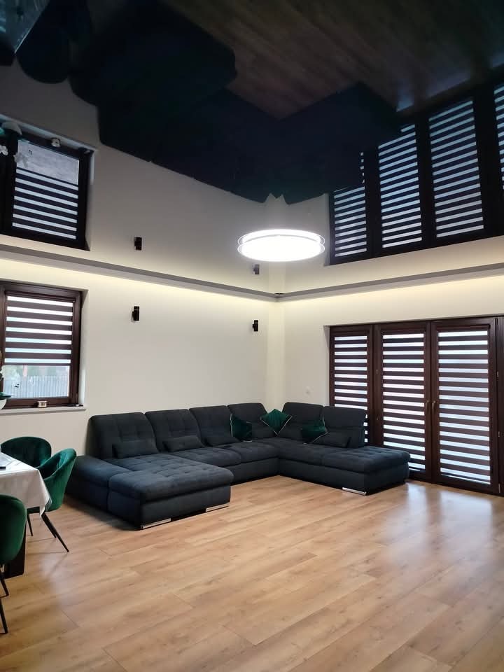 Nowoczesny salon z ciemną, błyszczącą podwieszaną powłoką sufitową, beżowymi ścianami, drewnianą podłogą i dużym, narożnym, ciemnoszarym wypoczynkiem z zielonymi poduszkami.