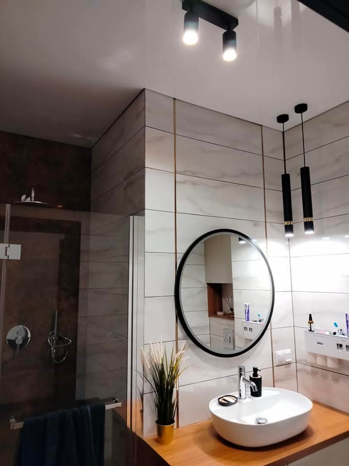 Nowoczesna łazienka z kabiną prysznicową, okrągłym lustrem i umywalką nablatową na drewnianym blacie, oświetlona czarnymi lampami sufitowymi.