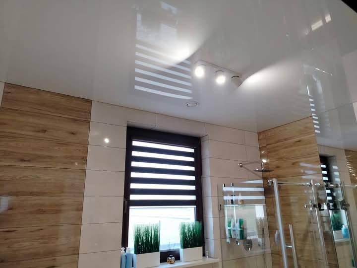 Nowoczesna łazienka z drewnopodobną okładziną ścienną, białymi płytkami, podwieszanym sufitem z oświetleniem punktowym i oknem z roletami dzień-noc.