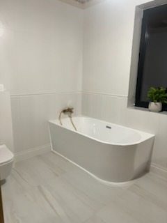 Biała, minimalistyczna łazienka z wolnostojącą wanną o zaokrąglonym kształcie, złotą armaturą i marmurową posadzką. W tle czarne okno z rośliną doniczkową. Widoczne delikatne listwy przypodłogowe.