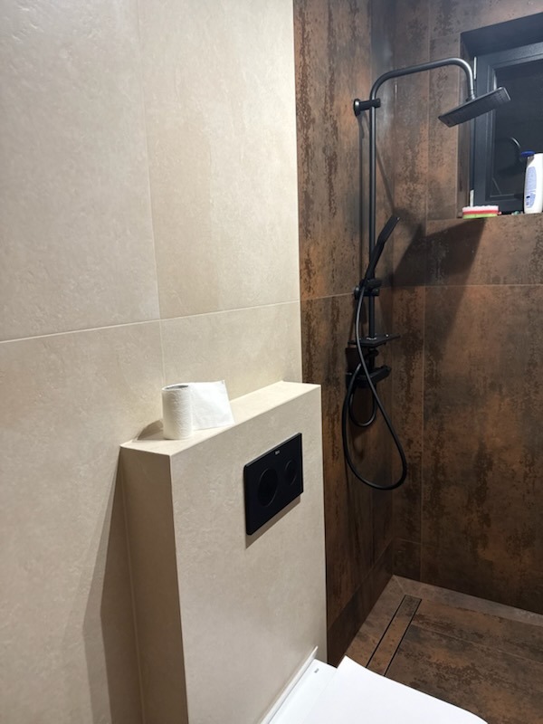 Nowoczesna łazienka z beżowymi i rdzawymi płytkami, czarną armaturą prysznicową i podtynkową spłuczką WC, na której leży rolka papieru toaletowego.