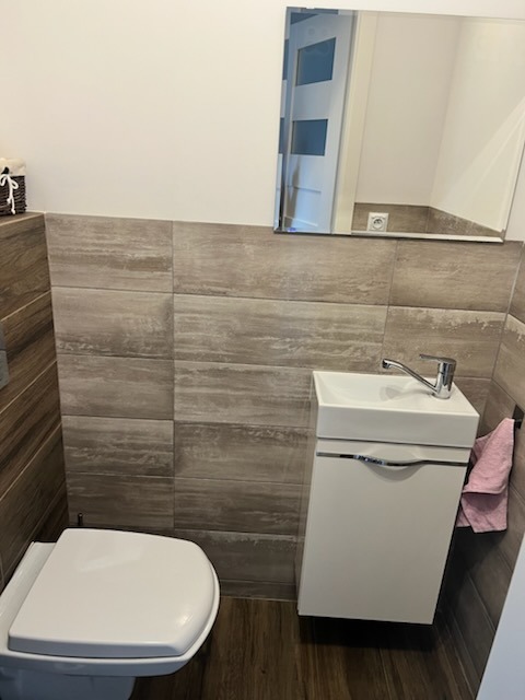 Nowoczesna, mała łazienka z umywalką nablatową na szafce, toaletą i lustrem, wykończona płytkami imitującymi drewno w odcieniach szarości i brązu.