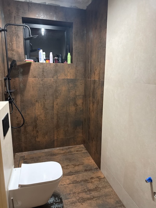 Nowoczesna łazienka z brązowymi i beżowymi płytkami, kabina prysznicowa typu walk-in z czarną armaturą, wisząca toaleta i okno z kosmetykami.