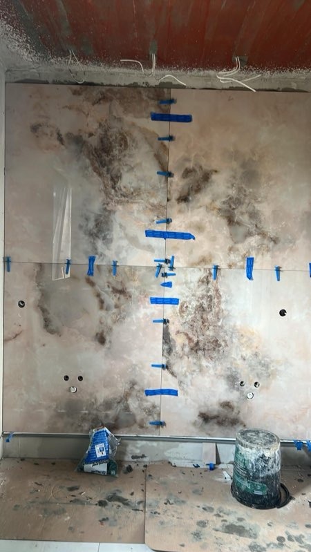 Montaż płytek imitujących marmur na ścianie, z użyciem niebieskiej taśmy maskującej do poziomowania i utrzymania odstępów. Widoczne puszki elektryczne i wiadro z zaprawą na podłodze.