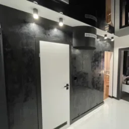 Nowoczesny korytarz z czarnymi ścianami o teksturze marmuru, białymi drzwiami z kratką wentylacyjną u dołu, błyszczącym czarnym sufitem podwieszanym z okrągłymi lampami i białymi płytkami podłogowymi.