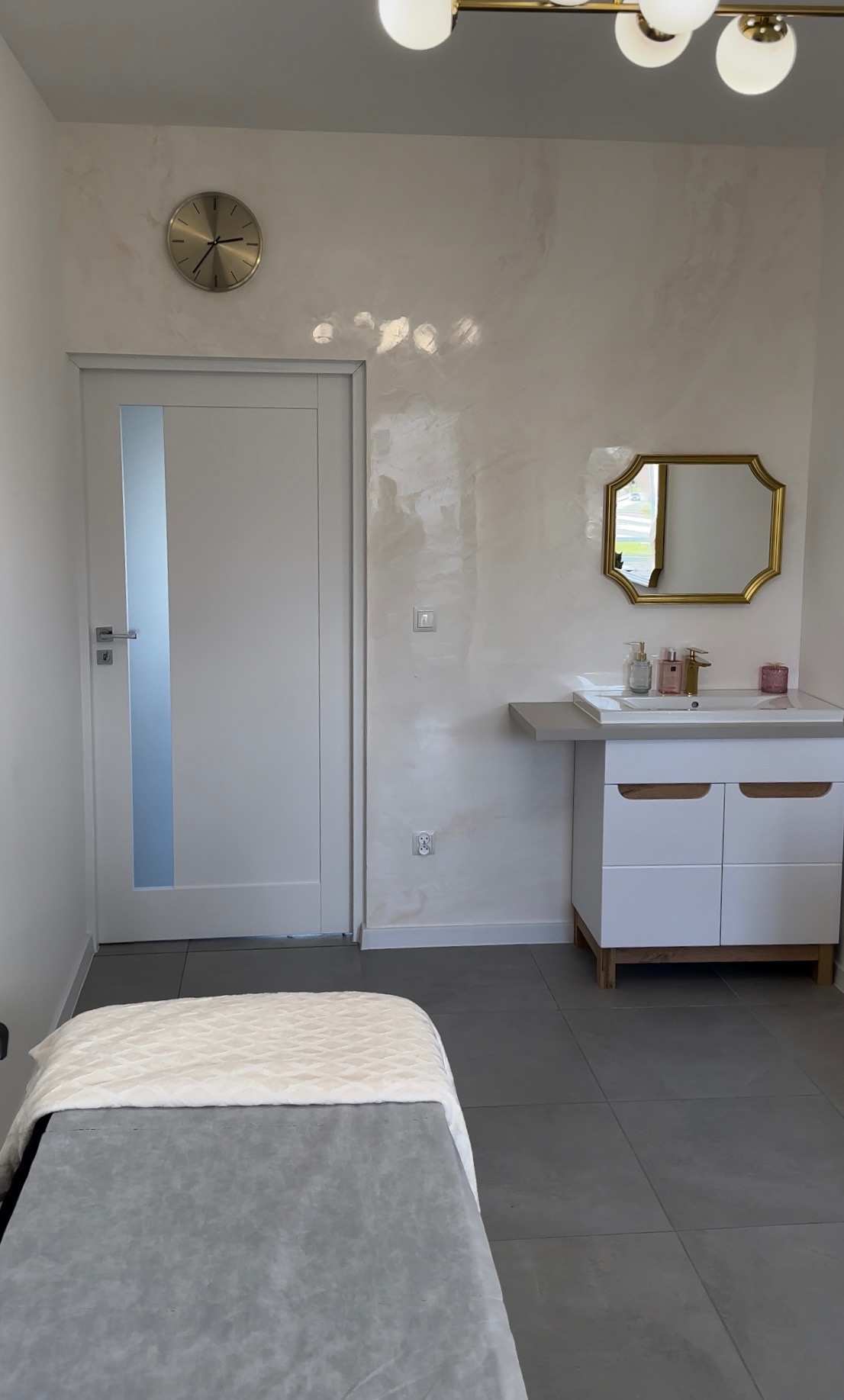 Wykończone wnętrze gabinetu kosmetycznego z dekoracyjną ścianą w odcieniach beżu, złotym zegarem i lustrem, białą szafką z umywalką oraz leżanką kosmetyczną z szarą tapicerką i kremowym pledem.