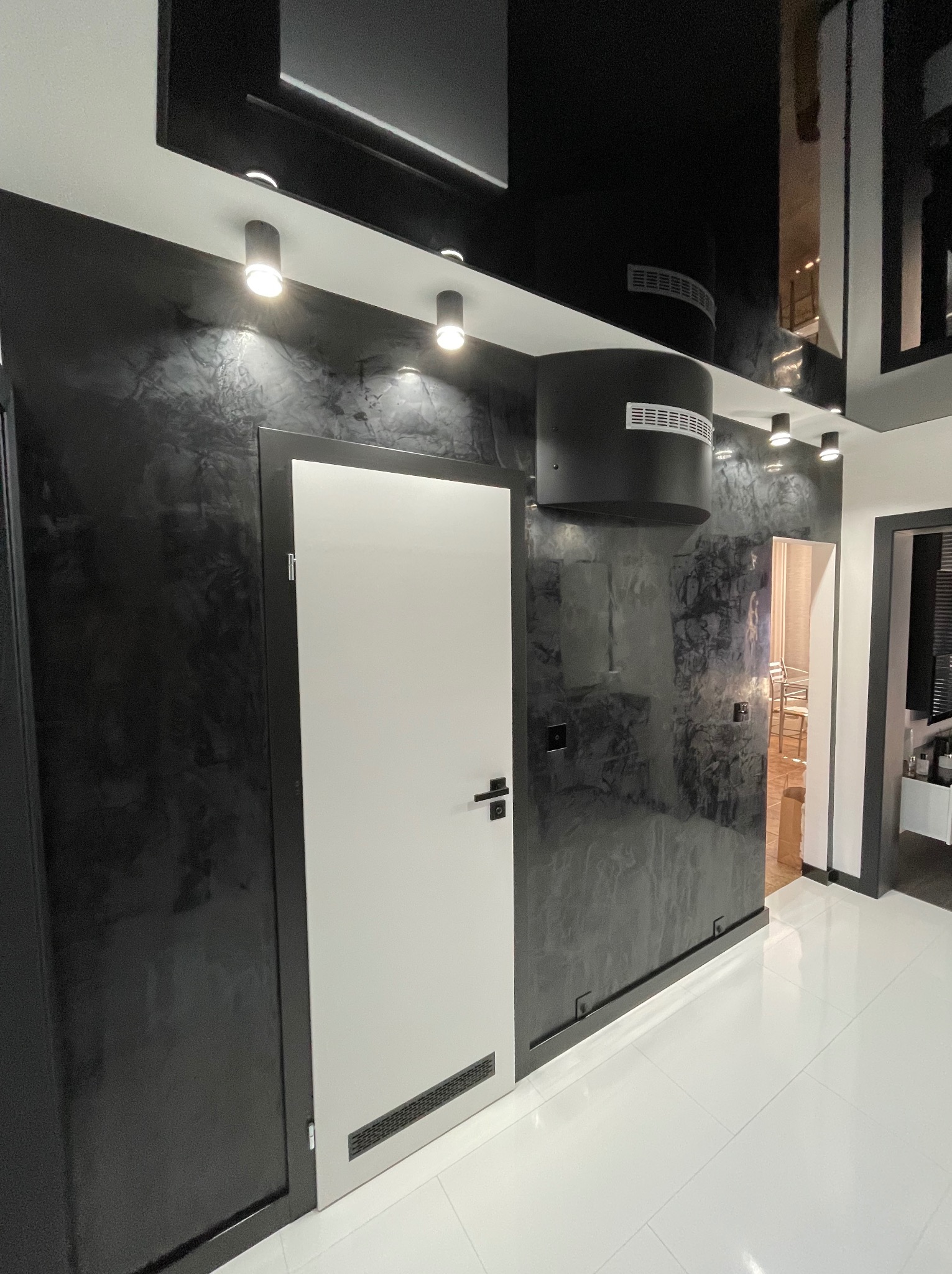 Nowoczesny korytarz z czarnymi ścianami o teksturze marmuru, białymi drzwiami z kratką wentylacyjną u dołu, błyszczącym czarnym sufitem podwieszanym z okrągłymi lampami i białymi płytkami podłogowymi.