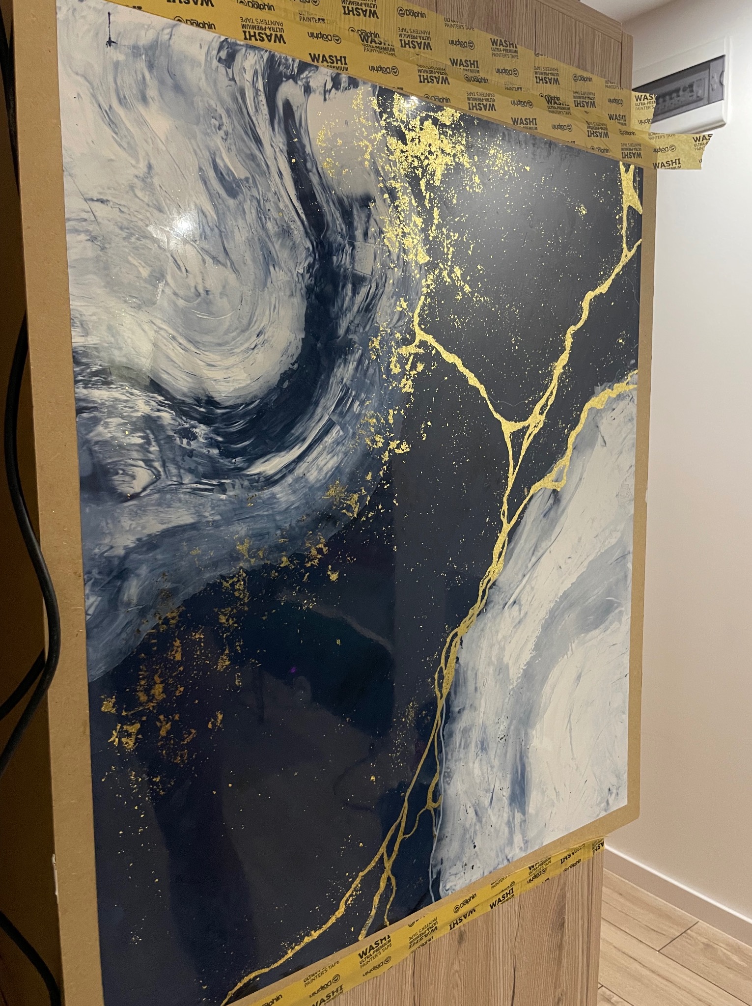 Abstrakcyjna kompozycja malarska w odcieniach bieli, granatu i złota, zabezpieczona taśmą malarską na drewnianej szafce, gotowa do montażu na ścianie.