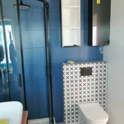 Nowoczesna łazienka z kabiną prysznicową typu walk-in z czarnymi profilami, toaletą podwieszaną z mozaiką na ścianie, niebieskimi płytkami ściennymi i białymi szafkami wiszącymi.