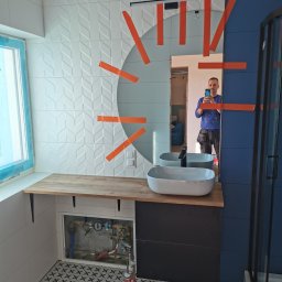 Witek - Łazienka w trakcie remontu: białe płytki z wzorem jodełki na ścianie, umywalka nablatowa na drewnianym blacie, czarna szafka pod umywalką, niebieska ściana, kabina prysznicowa w tle, widoczne rury...