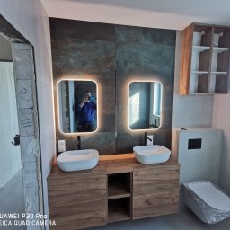 Witek - Nowoczesna łazienka w trakcie wykańczania: podwójna umywalka nablatowa na drewnianej szafce, podświetlane lustra, ciemne płytki na ścianie, częściowo zakryta toaleta w folii ochronnej, widoczne...