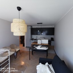 Witek - Wnętrze mieszkania w trakcie urządzania: podłoga w jodełkę, widoczne meble w paczkach, lampa wisząca z wikliny, kuchnia w tle.