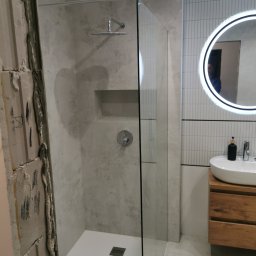 Witek - Wnętrze łazienki w trakcie remontu: widoczna kabina prysznicowa typu walk-in z deszczownicą i wnęką na kosmetyki, umywalka nablatowa na drewnianej szafce oraz fragment ściany w stanie surowym.