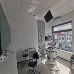 Nowoczesny gabinet stomatologiczny z fotelem, lampą i widokiem na budynek kantoru za oknem, białe ściany z dekoracyjnym panelem pionowym, osoba wykonująca prace wykończeniowe na podłodze.