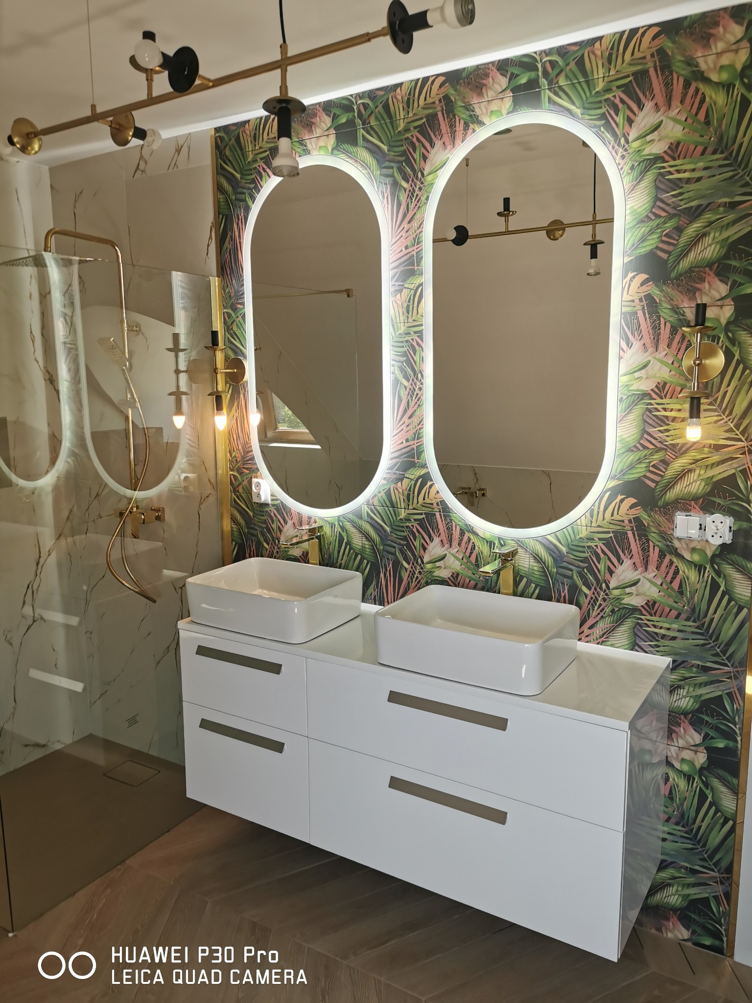 Nowoczesna łazienka z dwoma białymi umywalkami nablatowymi, złotą armaturą, podświetlanymi lustrami w kształcie owalu i ścianą w tropikalne liście, oświetlona złotymi lampami.