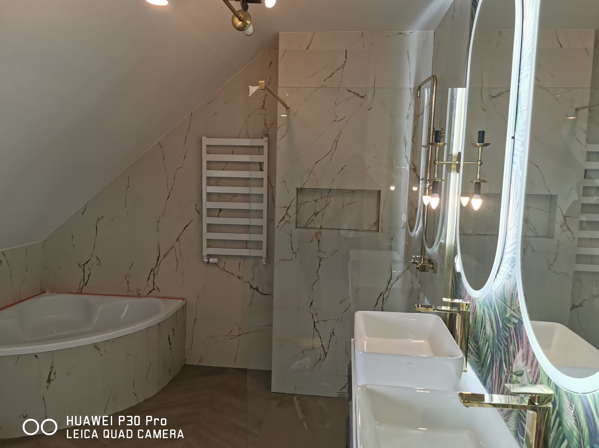 Nowoczesna łazienka na poddaszu z wanną narożną, marmurowymi płytkami ściennymi z brązowymi żyłami, grzejnikiem drabinkowym i podwójną umywalką z bateriami w kolorze złotym.