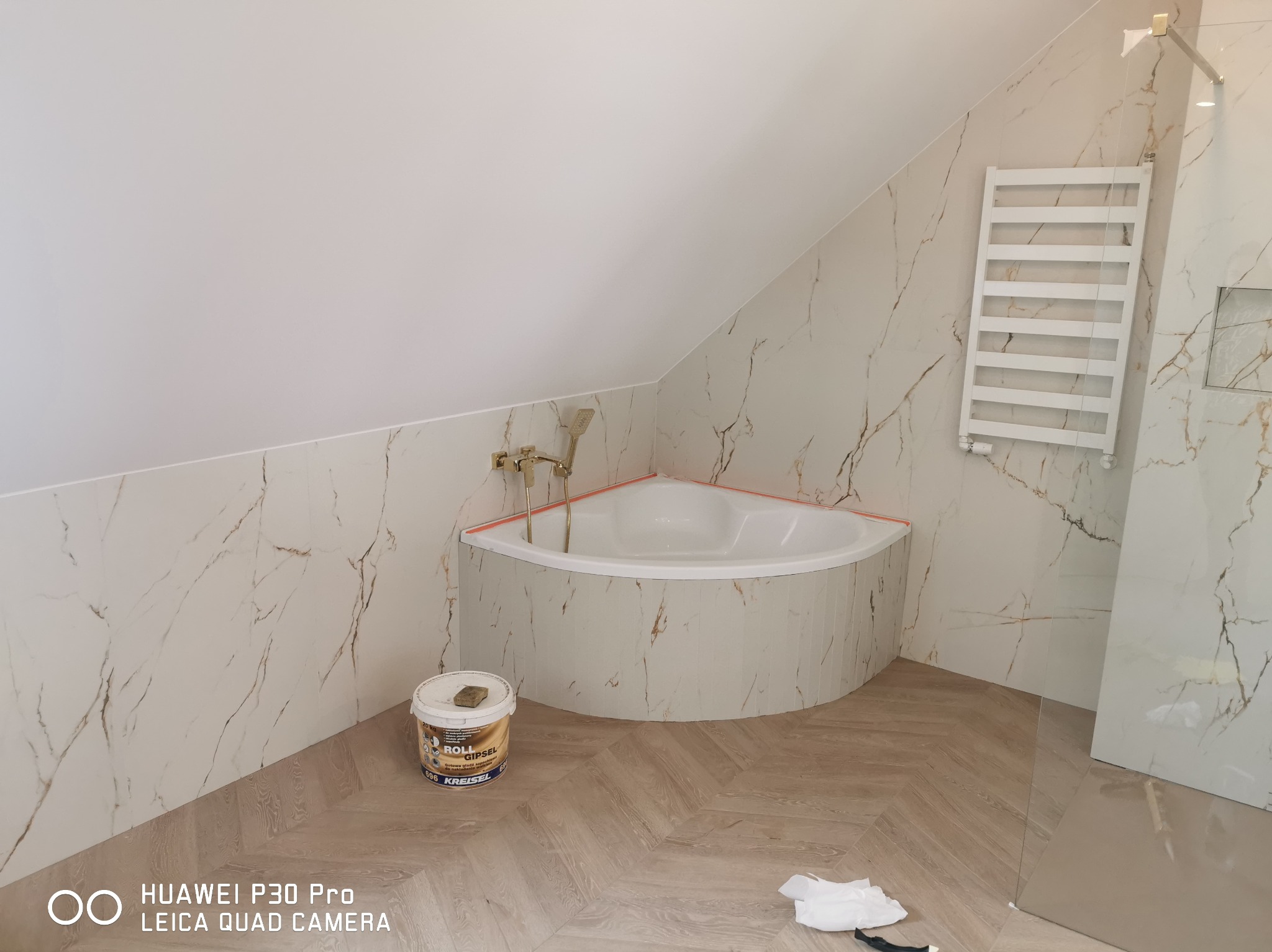 Wnętrze łazienki w trakcie wykończenia: narożna wanna obudowana płytkami imitującymi marmur, grzejnik drabinkowy, podłoga z paneli ułożonych w jodełkę, wiadro z gładzią szpachlową Kreisel na posadzce.