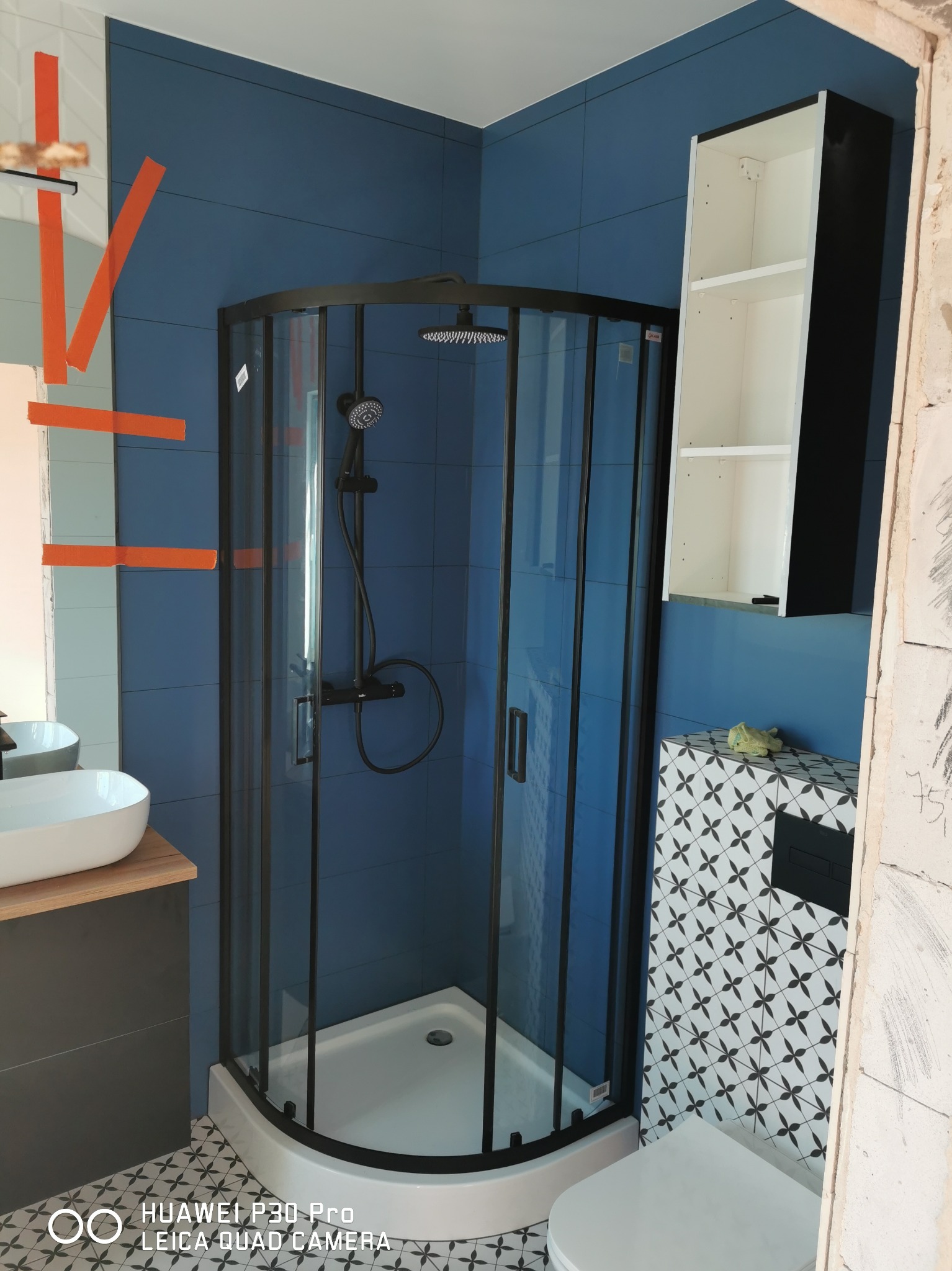 Niebieska łazienka z kabiną prysznicową z czarnymi ramami, umywalką nablatową na drewnianym blacie i toaletą z mozaiką na ścianie, częściowo wykończona.