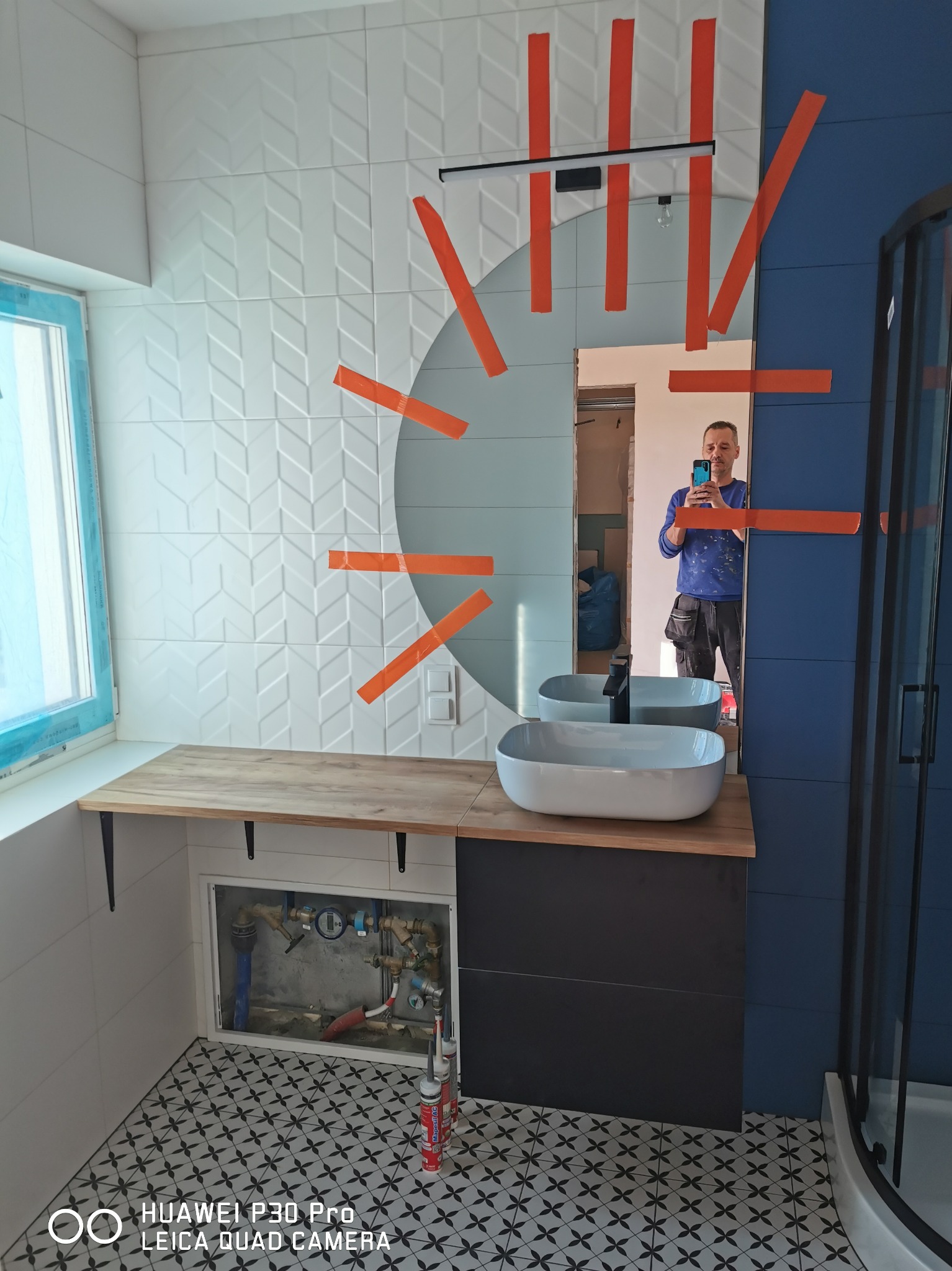 Łazienka w trakcie remontu: białe płytki z wzorem jodełki na ścianie, umywalka nablatowa na drewnianym blacie, czarna szafka pod umywalką, niebieska ściana, kabina prysznicowa w tle, widoczne rury...