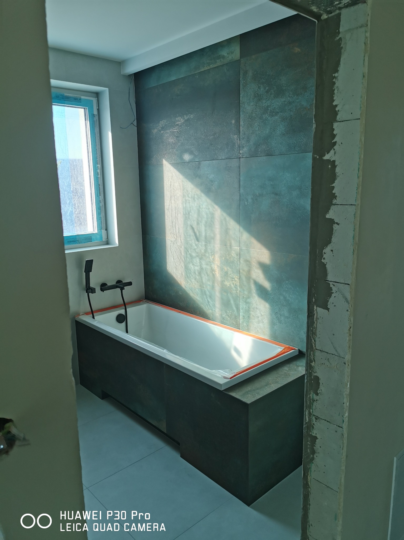 Nowoczesna łazienka w trakcie remontu: ciemne, duże płytki na ścianie, biała wanna z czarną armaturą, widoczne surowe wykończenie framugi drzwi, wpadające światło słoneczne przez okno.