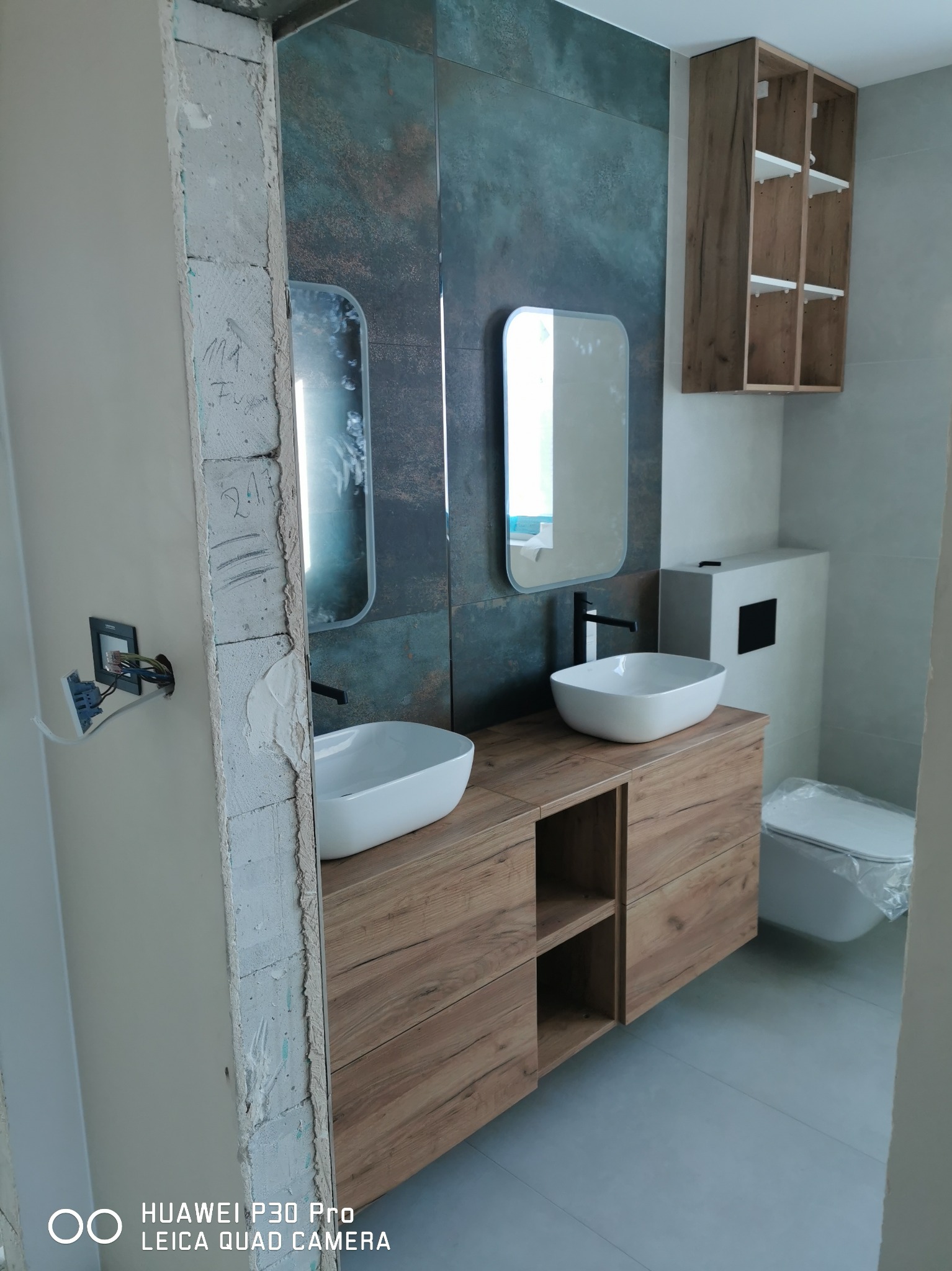 Łazienka w trakcie remontu: dwie umywalki nablatowe na drewnopodobnej szafce, lustra w ramie, płytki imitujące rdzę na ścianie, częściowo wykończona ściana z widoczną instalacją elektryczną.