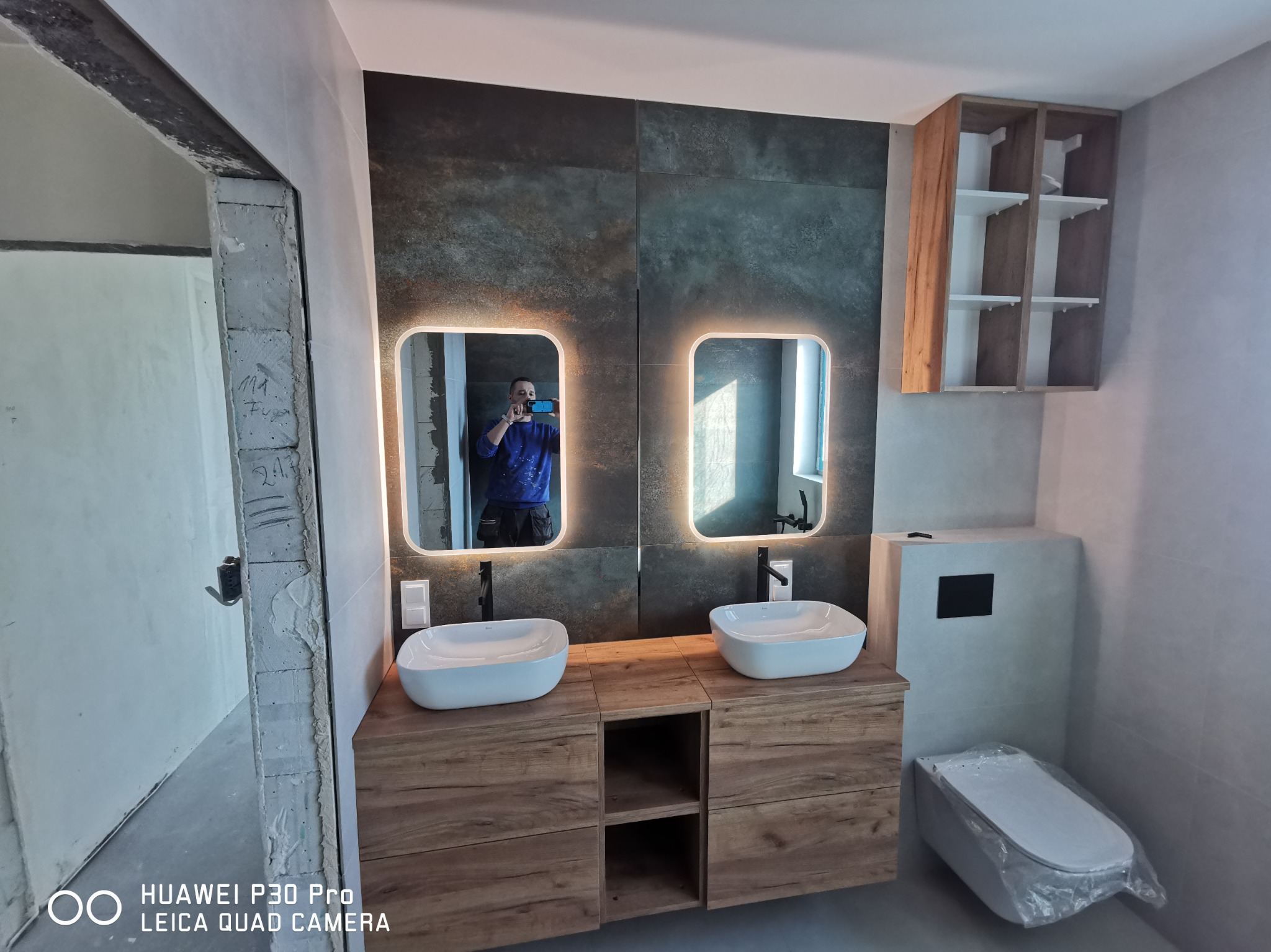 Nowoczesna łazienka w trakcie wykańczania: podwójna umywalka nablatowa na drewnianej szafce, podświetlane lustra, ciemne płytki na ścianie, częściowo zakryta toaleta w folii ochronnej, widoczne...