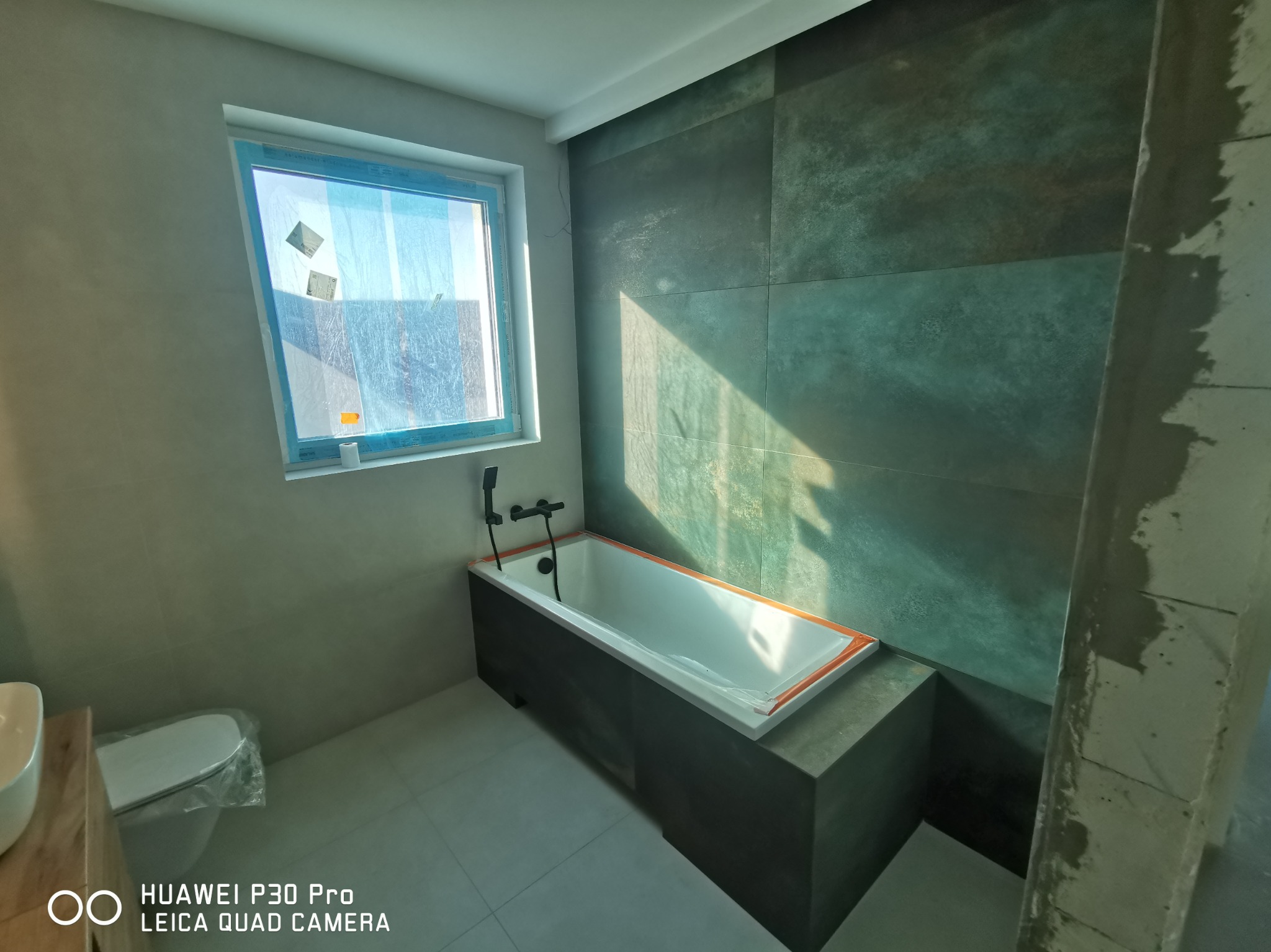 Wnętrze łazienki w trakcie wykończenia: biała wanna z czarną armaturą, szare płytki ścienne i podłogowe, okno z folią ochronną, częściowo zamontowana toaleta z folią ochronną, widoczny surowy filar.