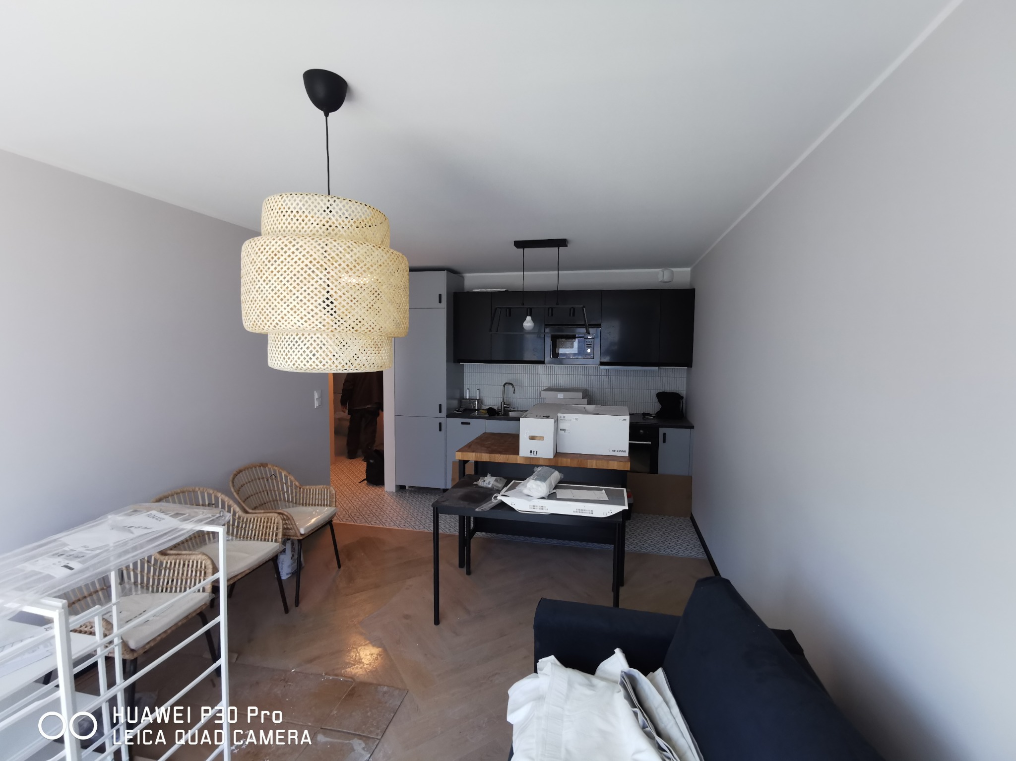 Wnętrze mieszkania w trakcie urządzania: podłoga w jodełkę, widoczne meble w paczkach, lampa wisząca z wikliny, kuchnia w tle.