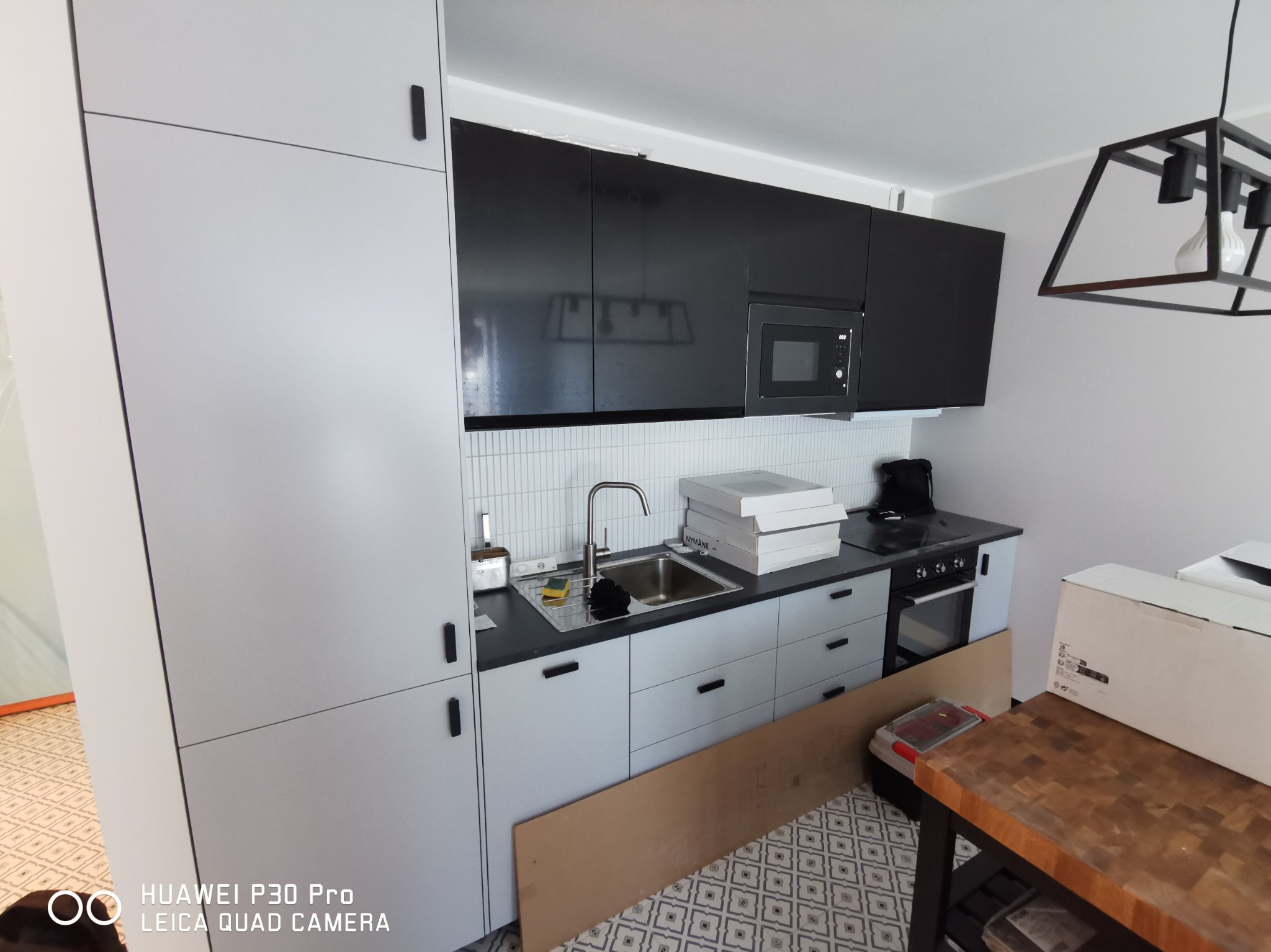 Nowoczesna kuchnia w trakcie montażu: szare szafki dolne i białe górne, czarne fronty szafek wiszących i mikrofalówka, biała mozaika na ścianie, kartony z wyposażeniem na blacie, deska...