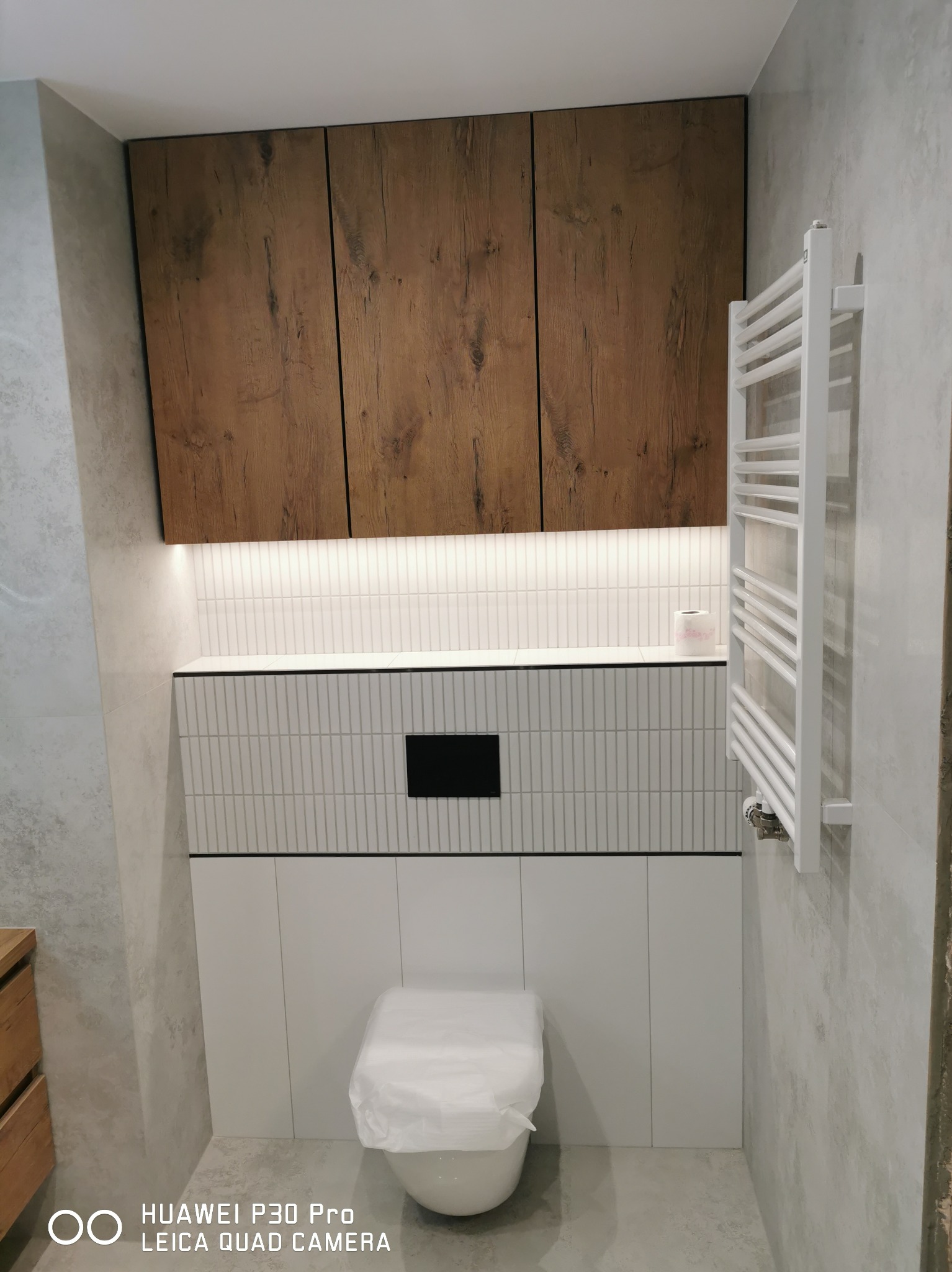 Nowoczesna łazienka z drewnianymi szafkami wiszącymi, podświetlanymi od dołu, pionowymi białymi płytkami i grzejnikiem drabinkowym, z toaletą zakrytą folią ochronną.