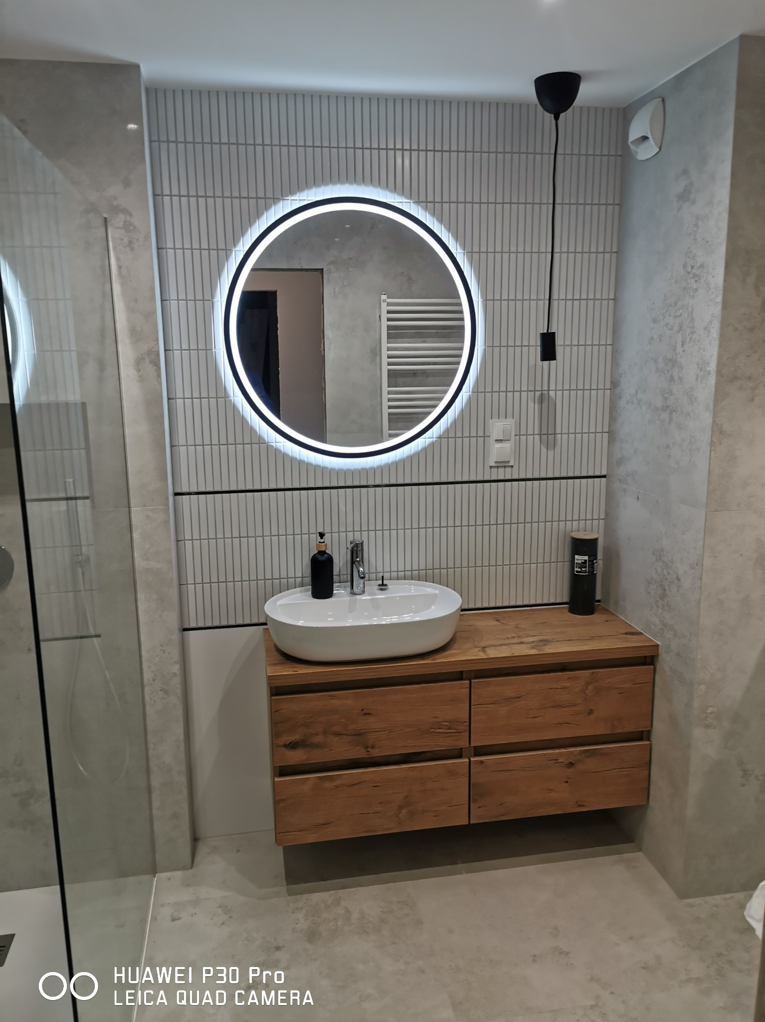 Nowoczesna łazienka w Starkowej Hucie z okrągłym lustrem LED, umywalką nablatową na drewnianej szafce i pionowymi białymi płytkami na ścianie.