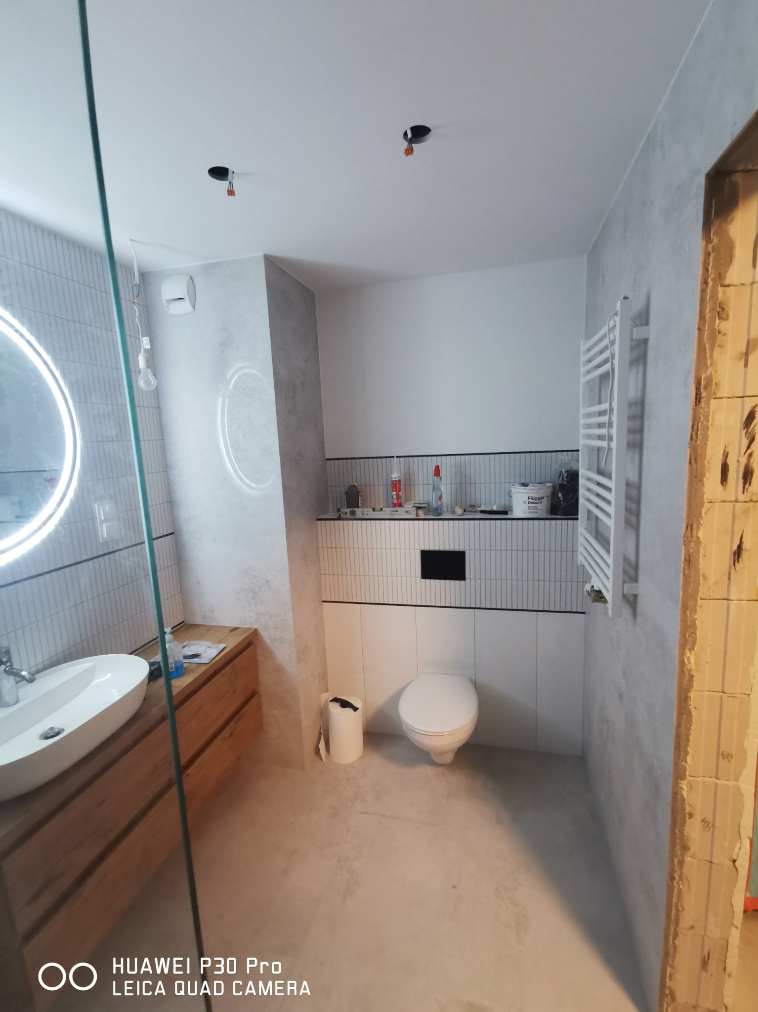 Wnętrze łazienki w trakcie remontu, widoczne płytki na ścianach, umywalka z drewnianą szafką, toaleta oraz grzejnik, surowy otwór drzwiowy po prawej stronie.