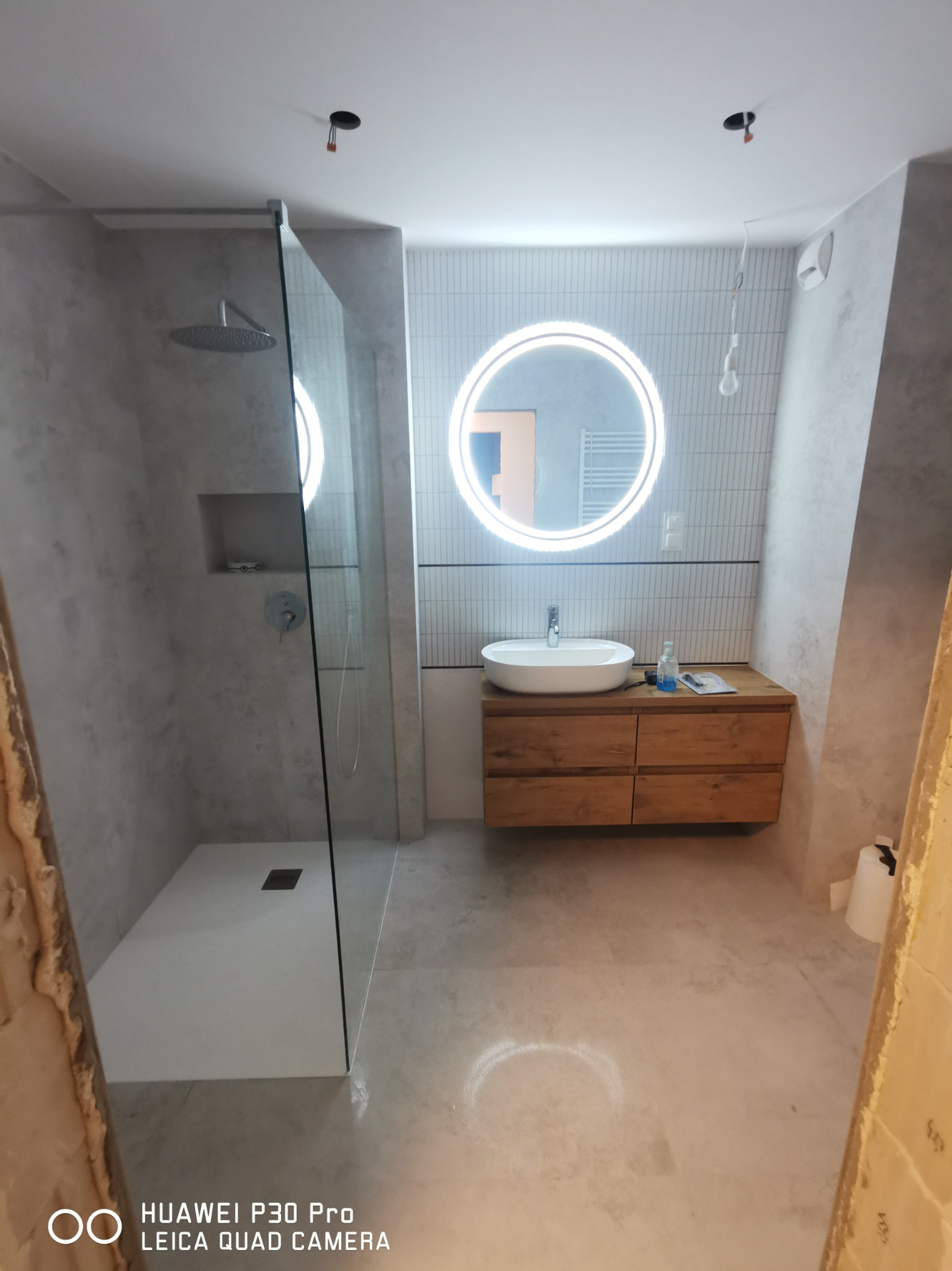 Nowoczesna łazienka w trakcie wykończenia: kabina prysznicowa typu walk-in z deszczownicą, okrągłe lustro z podświetleniem LED nad drewnianą szafką z umywalką, surowe ściany z efektem betonu...