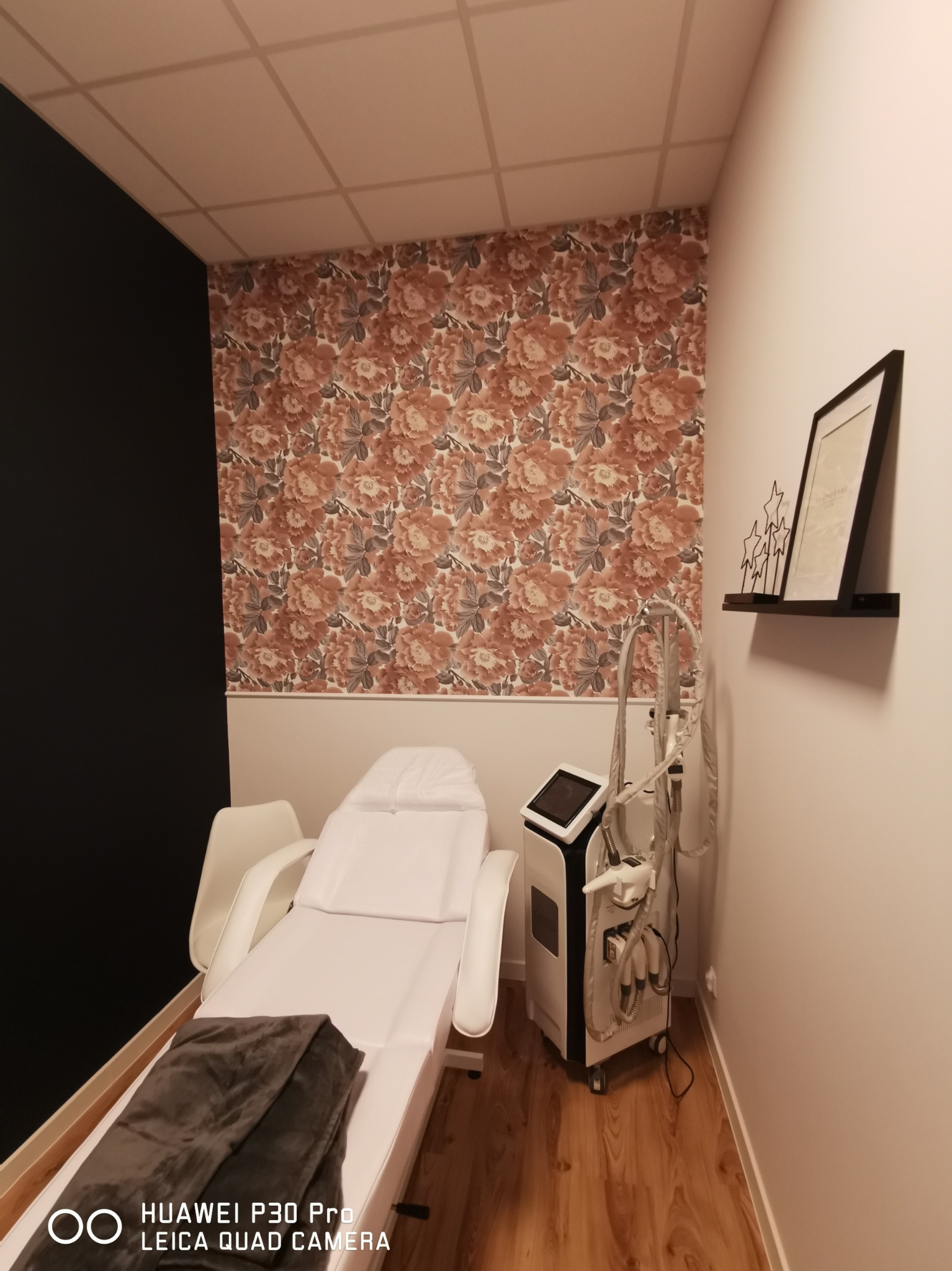 Gabinet kosmetyczny z białym fotelem zabiegowym, urządzeniem do modelowania sylwetki i ścianą w kwiatowy wzór