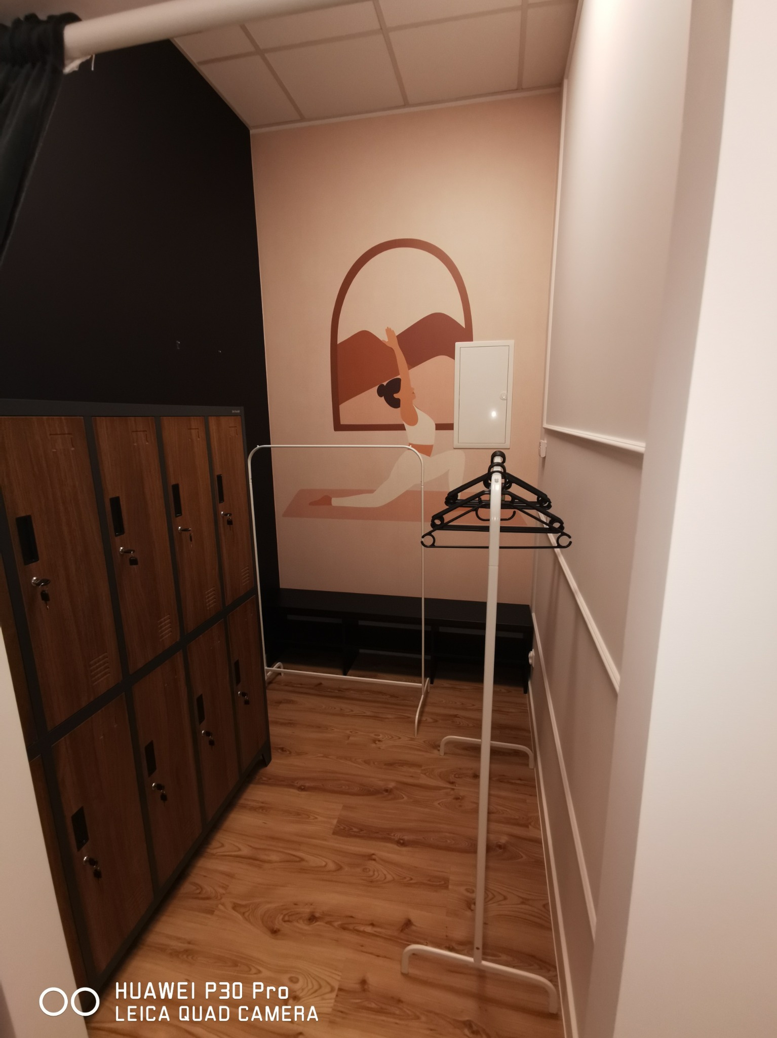 Szczegółowe ujęcie szatni z drewnianymi szafkami na ubrania, wieszakiem na ubrania oraz malowidłem na ścianie przedstawiającym postać w pozycji jogi, na podłodze panele.