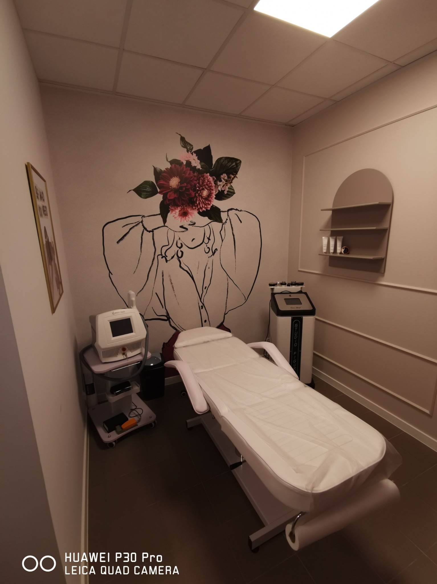 Jasne wnętrze gabinetu kosmetycznego z białym łóżkiem zabiegowym, aparaturą kosmetyczną i dekoracyjną grafiką na ścianie