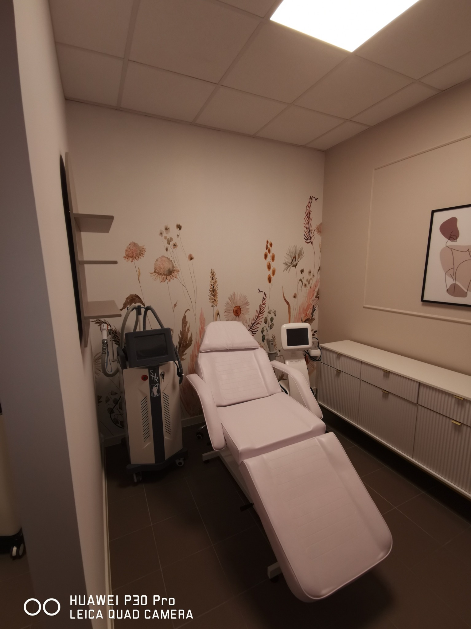 Jasne wnętrze gabinetu kosmetycznego z beżowymi ścianami, fototapetą z motywem kwiatowym, białym fotelem kosmetycznym i urządzeniem do zabiegów laserowych z wyświetlaczem.