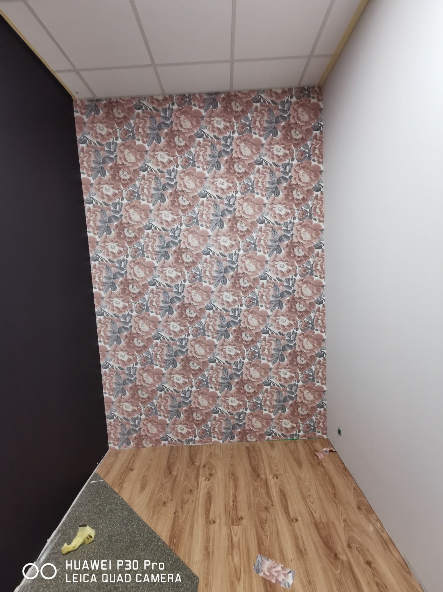Pokój po remoncie: ściana z tapetą w kwiaty, panele podłogowe imitujące drewno, czarna ściana po lewej i biała ściana po prawej stronie, widoczne fragmenty sufitu kasetonowego.