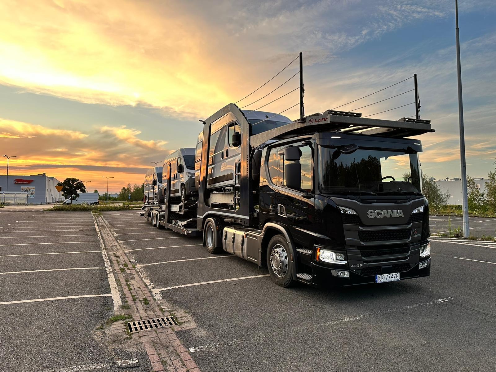 Czarna laweta Scania 450P z trzema busami na platformie, zaparkowana na parkingu z widokiem na zachodzące słońce.