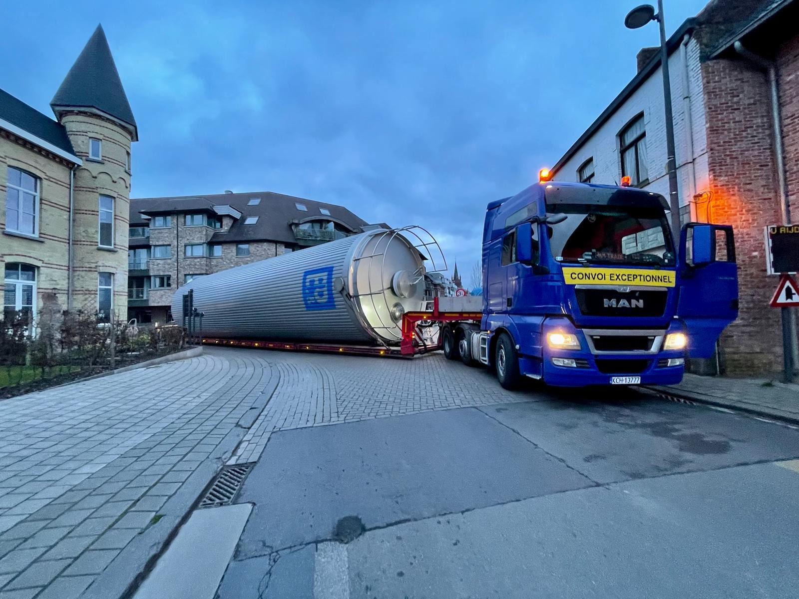 Niebieski ciągnik MAN z naczepą przewozi duży, poziomy zbiornik w zabudowie miejskiej, na boku zbiornika widoczne logo.