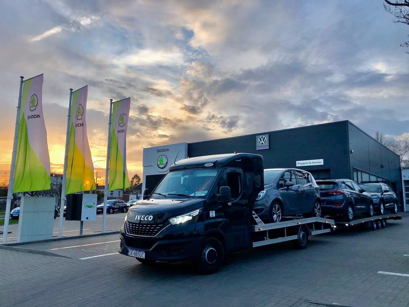 Czarna laweta Iveco z trzema samochodami osobowymi na platformie, zaparkowana przed salonem Skody i Volkswagena o zmierzchu.