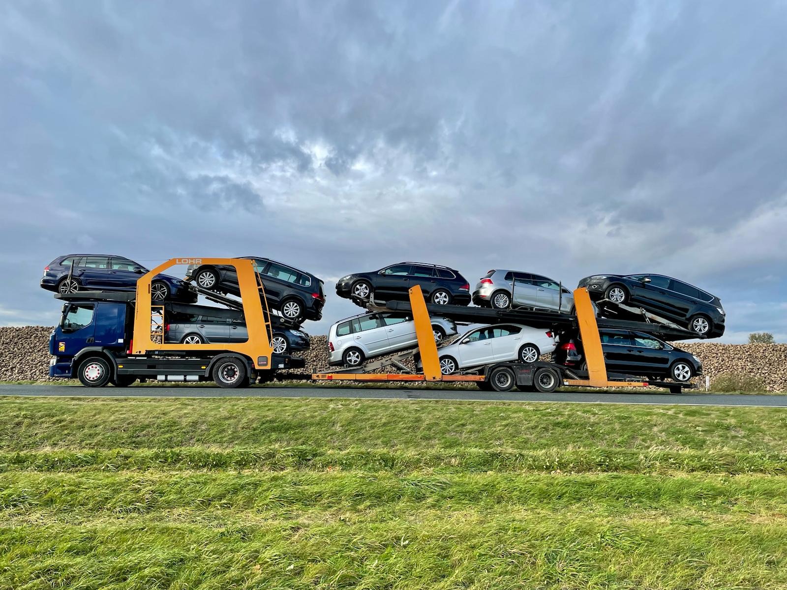 Niebieska ciężarówka z pomarańczowym stelażem marki Lohr przewozi kilka samochodów osobowych różnej marki i koloru na tle pochmurnego nieba i zielonego pola.